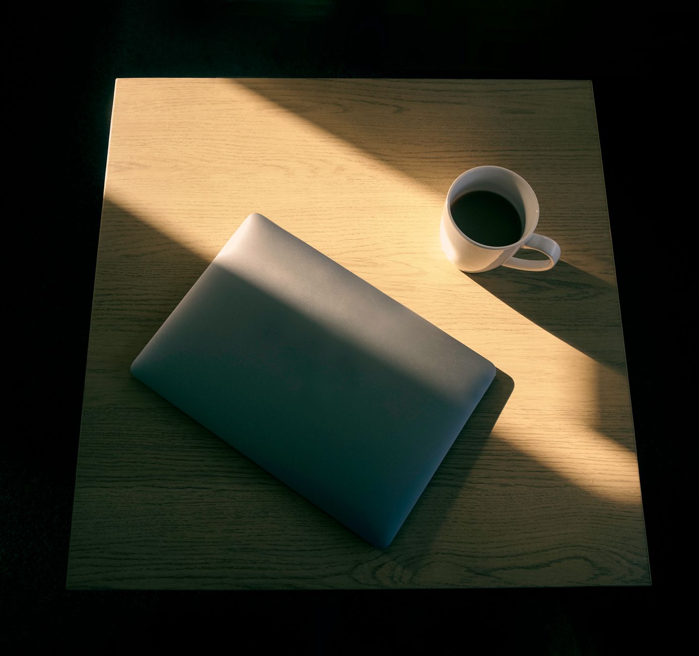 Minimalistischer Arbeitsplatz mit Laptop und einer Tasse Kaffee auf einem Holztisch im Licht-Schatten-Spiel.