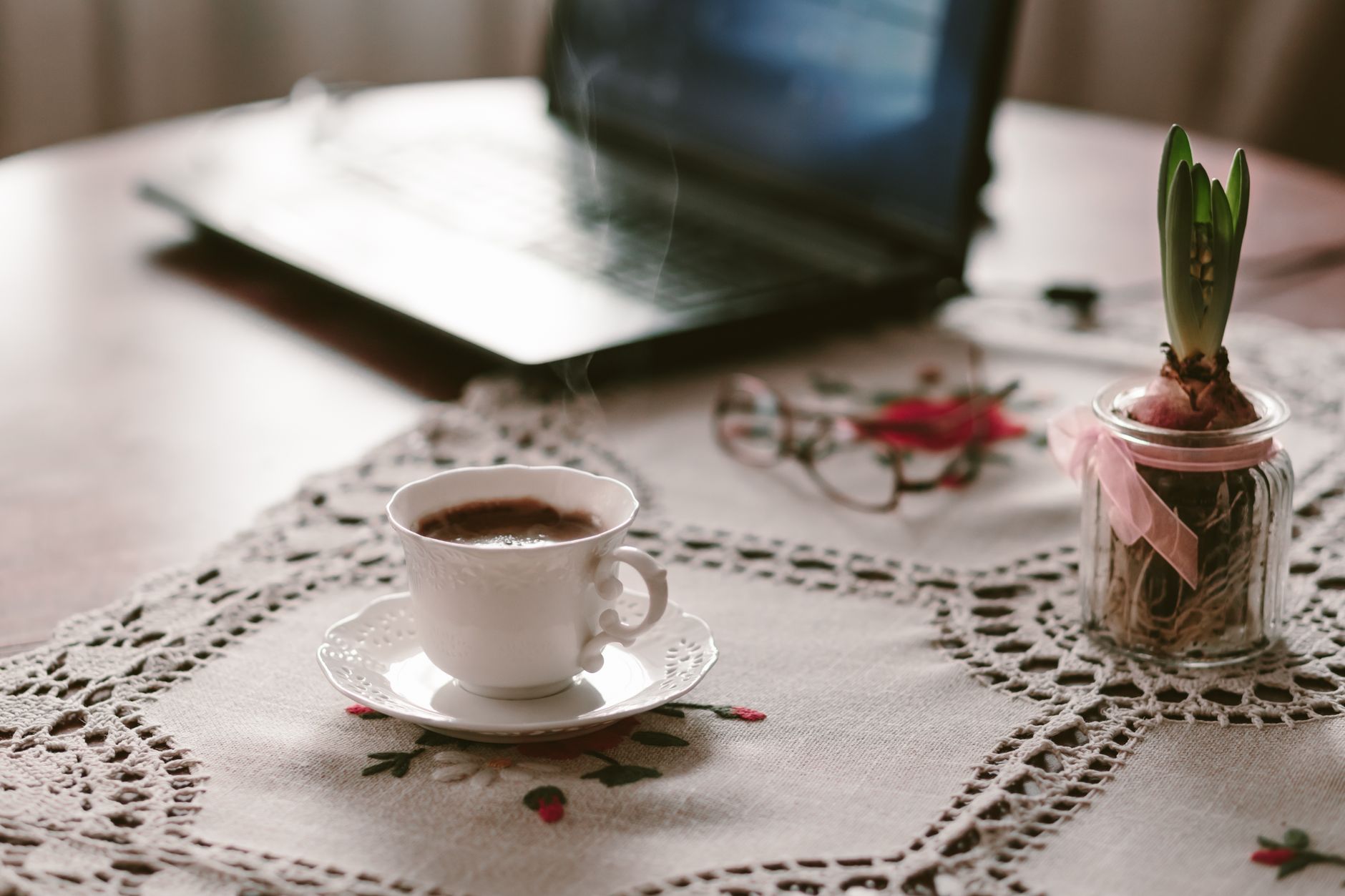 Warmer, einladender Arbeitsplatz mit einer dampfenden Tasse Kaffee auf einer Spitzendecke neben einem Laptop.